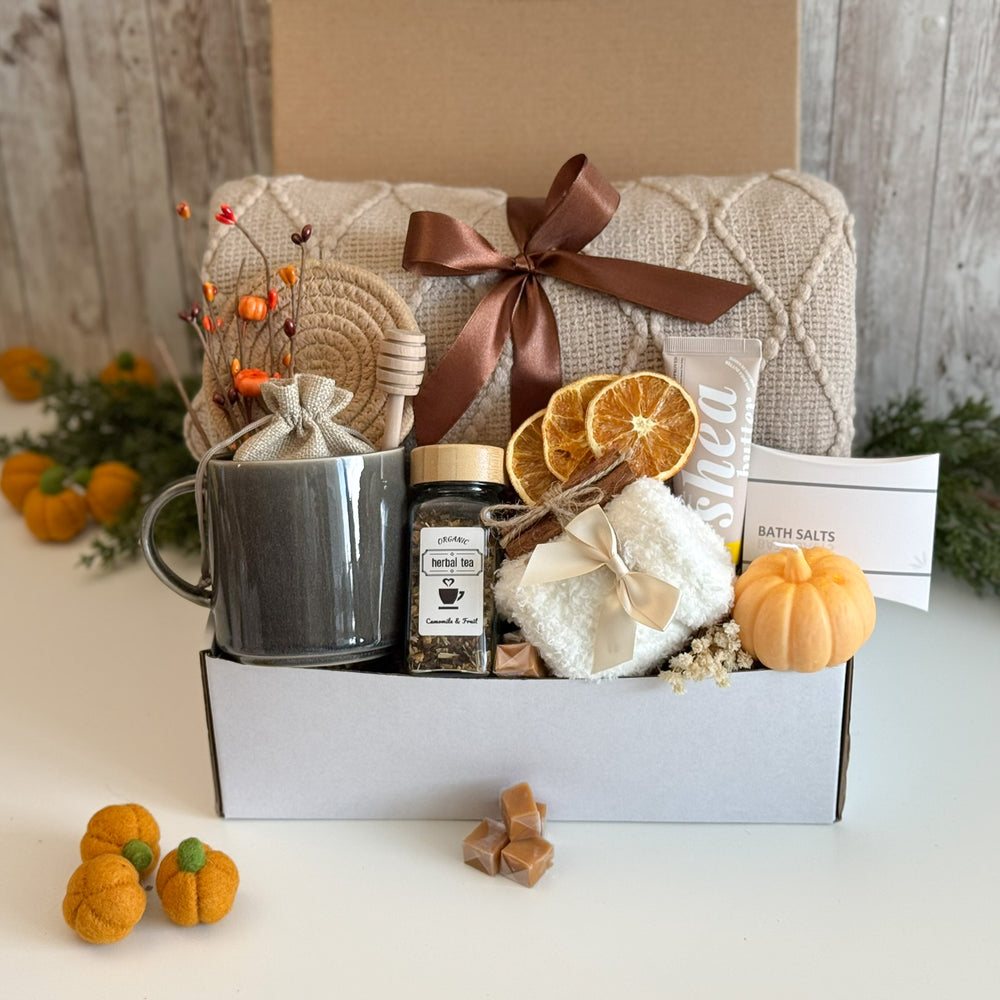 A curated gift box set featuring a gray ceramic mug with dried orange branches, a jar of herbal tea, bath salts, a white bath towel, and small orange pumpkins, arranged in a white box with a beige quilted blanket and brown ribbon in the background.