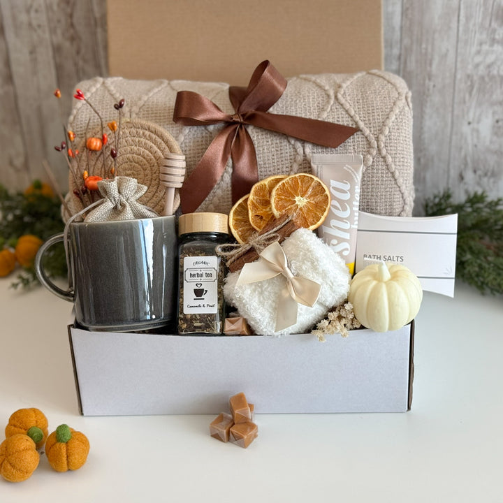 A curated gift box set on a white surface featuring a gray ceramic mug, a jar of herbal tea, a white towel, dried orange slices, a small cream pumpkin, a brown ribbon, and a card labeled 'PATH SALTS'.