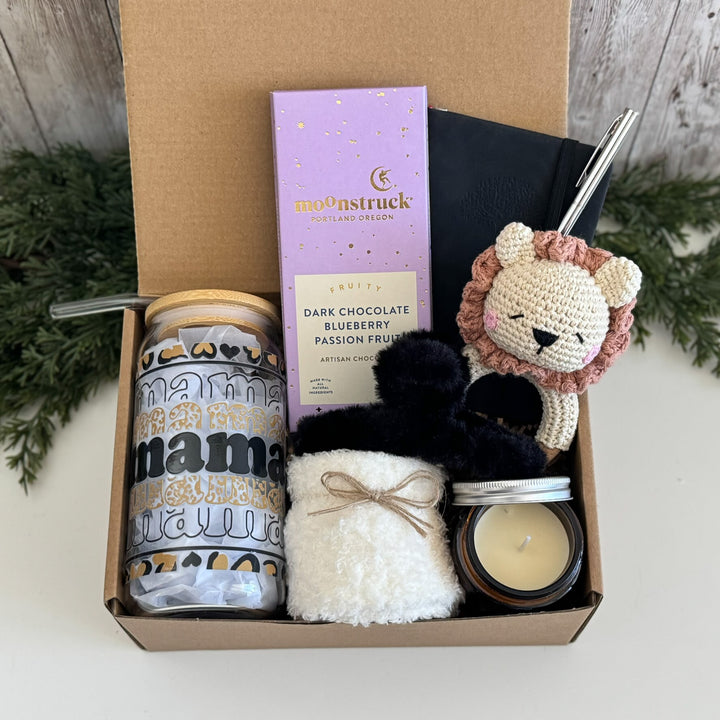 A curated gift box set containing a patterned tumbler, a Moonstruck chocolate bar, a crocheted lion plush, a black pom-pom, a white fluffy item tied with twine, and a candle in a cardboard box.
