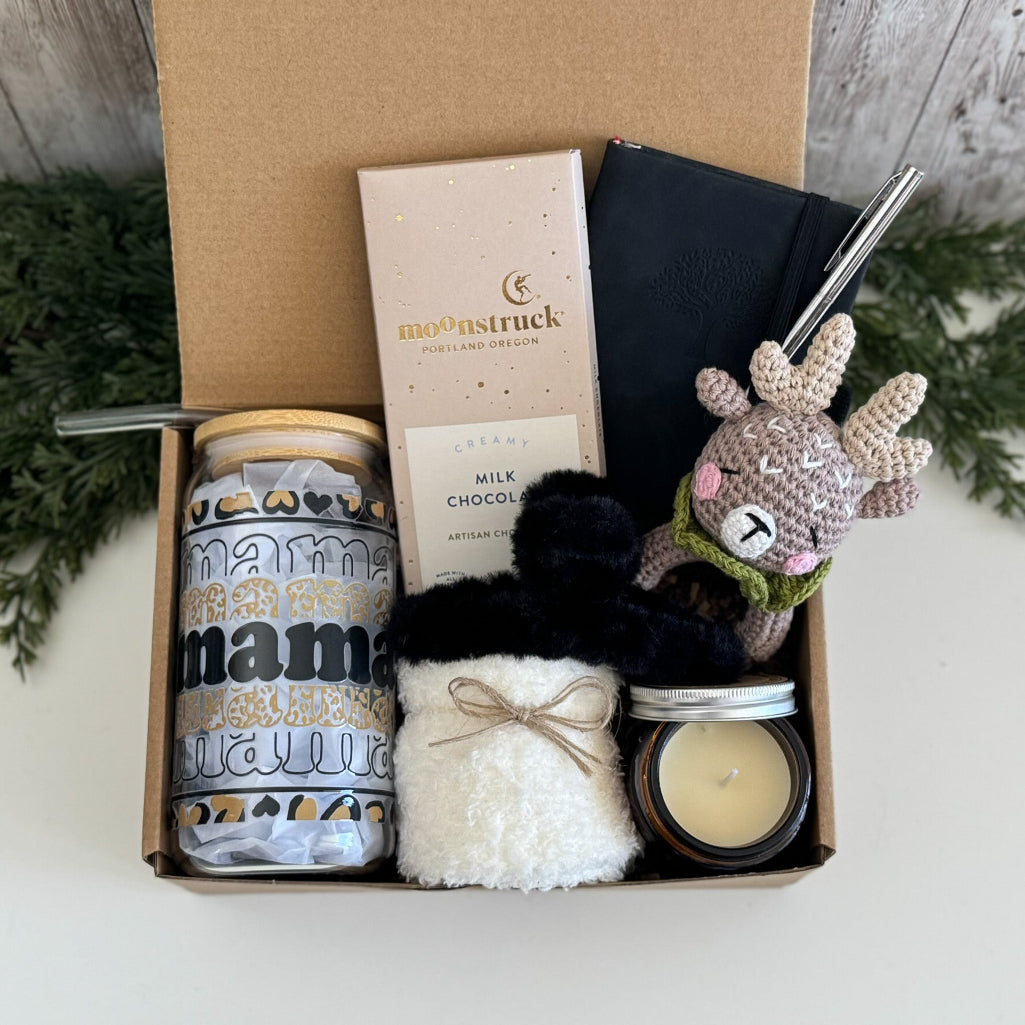A curated gift box set containing a 'mama natural' tumbler, a Moonstruck milk chocolate bar, a black notebook with a silver pen, a white fluffy pouch, a candle, and a crocheted reindeer toy, all arranged inside an open brown cardboard box.