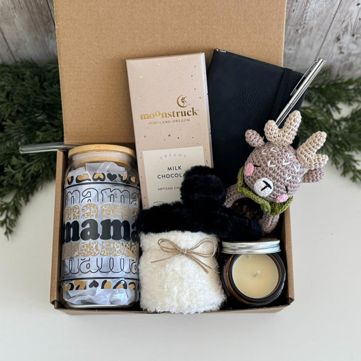 A curated gift box set containing a 'mama natural' tumbler, a Moonstruck milk chocolate bar, a black notebook with a silver pen, a white fluffy pouch, a candle, and a crocheted reindeer toy, all arranged inside an open brown cardboard box.