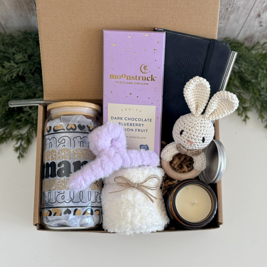 A curated gift box set containing a lavender Moonstruck chocolate bar, a white crochet bunny plush toy, a white fuzzy pouch tied with twine, a patterned tumbler, a candle, and a notebook, all arranged inside an open cardboard box.