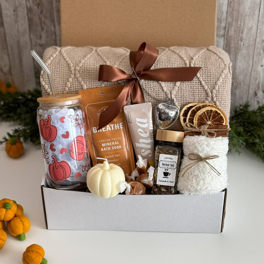 A curated gift box set displayed on a white surface with a rustic wood backdrop. The set includes a knitted beige towel tied with a brown ribbon, a patterned glass jar with a metal straw, a brown bath star, a white bath soak bag, a small white pumpkin, a jar of honey, dried orange slices, and a bundle of white bath towels.