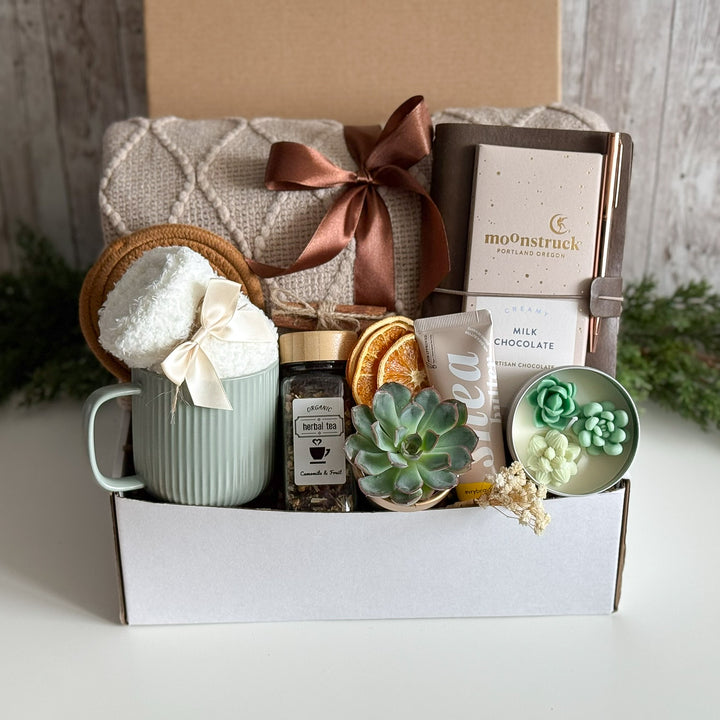 A curated gift box set on a white surface against a rustic wood backdrop. The set includes a mint green ribbed mug with a white towel and cream ribbon, a brown knit blanket tied with a satin ribbon, a notebook from moonstruck, a milk chocolate bar, a jar of herbal tea, dried orange slices, a small succulent, and a dish of green flower-shaped candies.