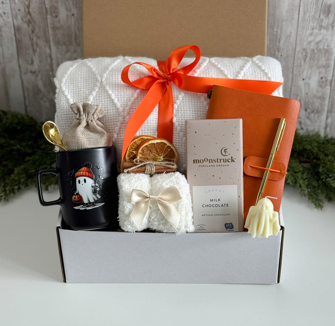 A curated gift box set on a white surface with a rustic wood backdrop and greenery. The set includes a white quilted blanket tied with an orange ribbon, a black mug with a ghost graphic and gold spoon, a small cream towel with a bow, dried orange slices, a milk chocolate bar, an orange notebook with a gold pen, and a small cream ghost-shaped item.