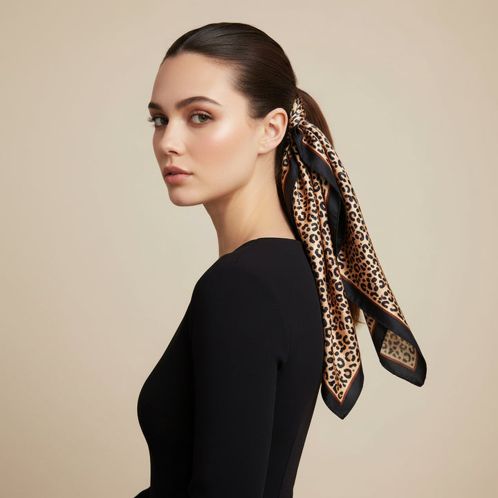 A woman with her hair pulled back wears a leopard-print silk scarf tied around her low ponytail, paired with a black long-sleeve top against a solid beige background.