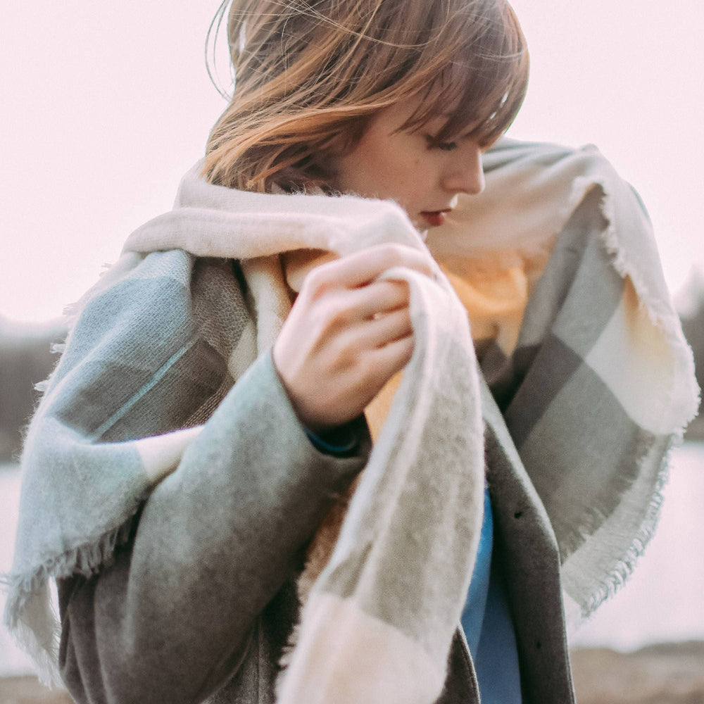 A woman outdoors near a body of water wearing a soft plaid scarf in muted gray, cream, and beige tones over a dark coat.