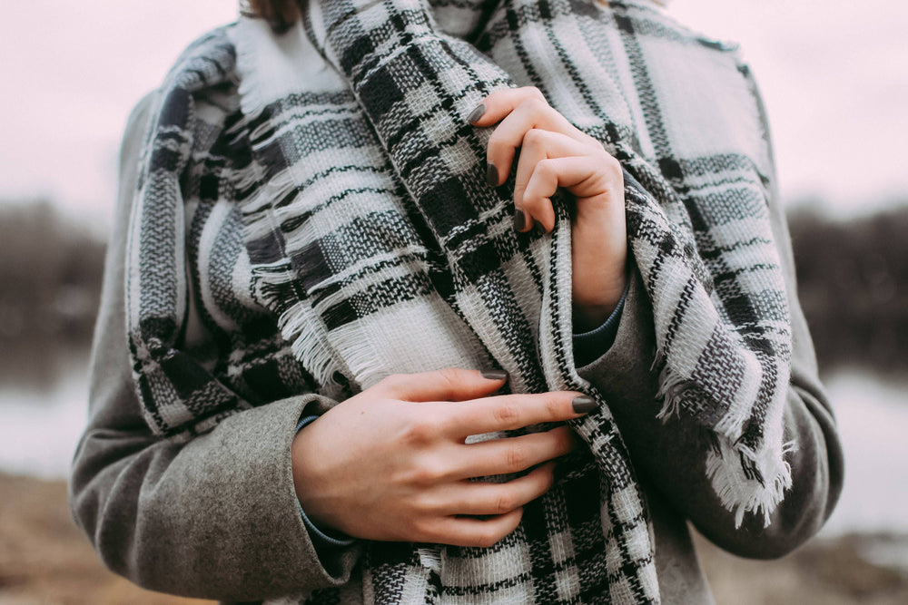 A person holding a black and white plaid blanket scarf with fringed ends, wrapped around their shoulders.