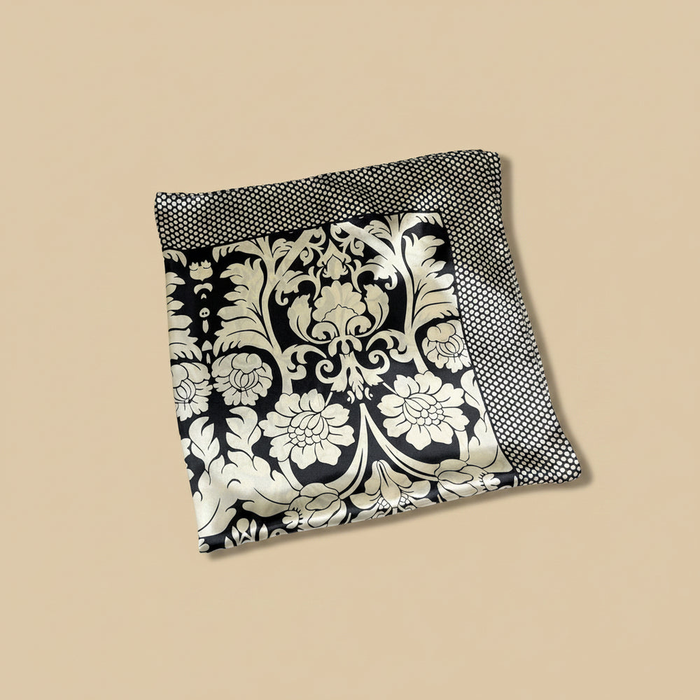 A folded square scarf with a black and beige damask floral pattern and a black-and-beige polka-dot border, displayed on a solid beige background.