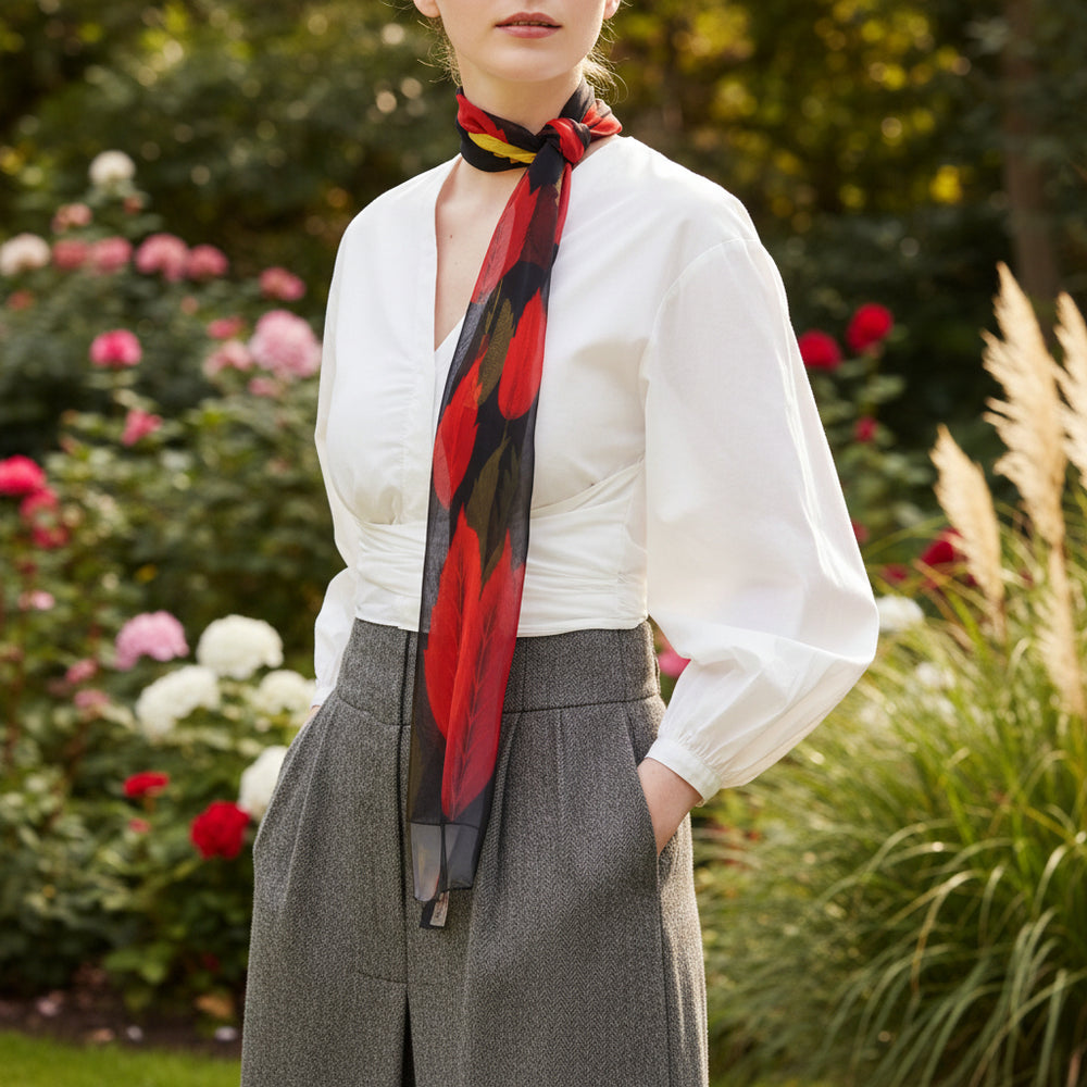 A woman wearing a white blouse and gray trousers models a sheer black scarf with large red and orange floral motifs in an outdoor garden setting.
