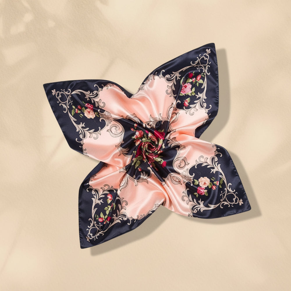 A square silk scarf with a luxurious satin sheen, featuring a design of alternating peach-pink and black sections decorated with gold baroque scrolls and pink floral motifs, arranged in a bow on a white marble background.