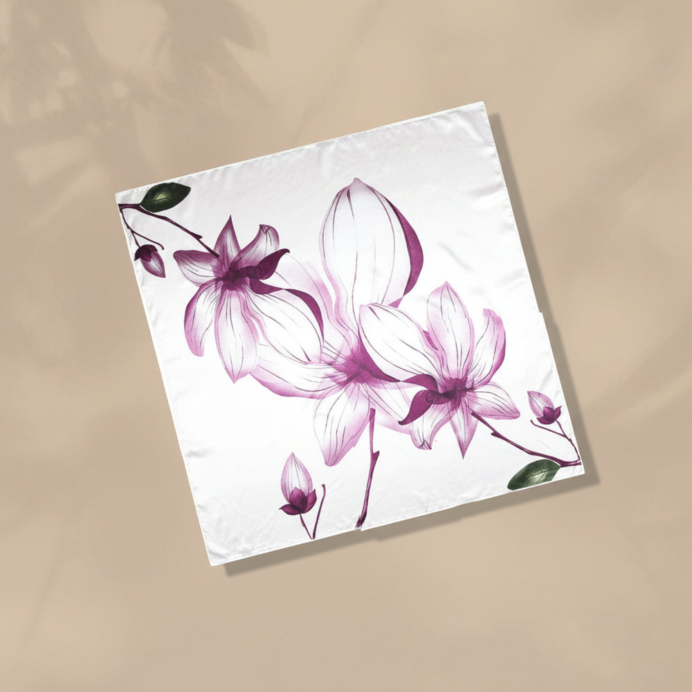 A square white fabric napkin or small tablecloth featuring a watercolor-style floral print in shades of purple, pink, and green, displayed diagonally on a beige background.