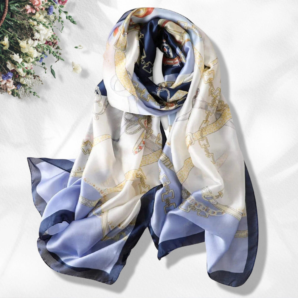 A navy and white silk scarf with floral and geometric patterns, draped elegantly on a white surface with a small floral arrangement in the background.